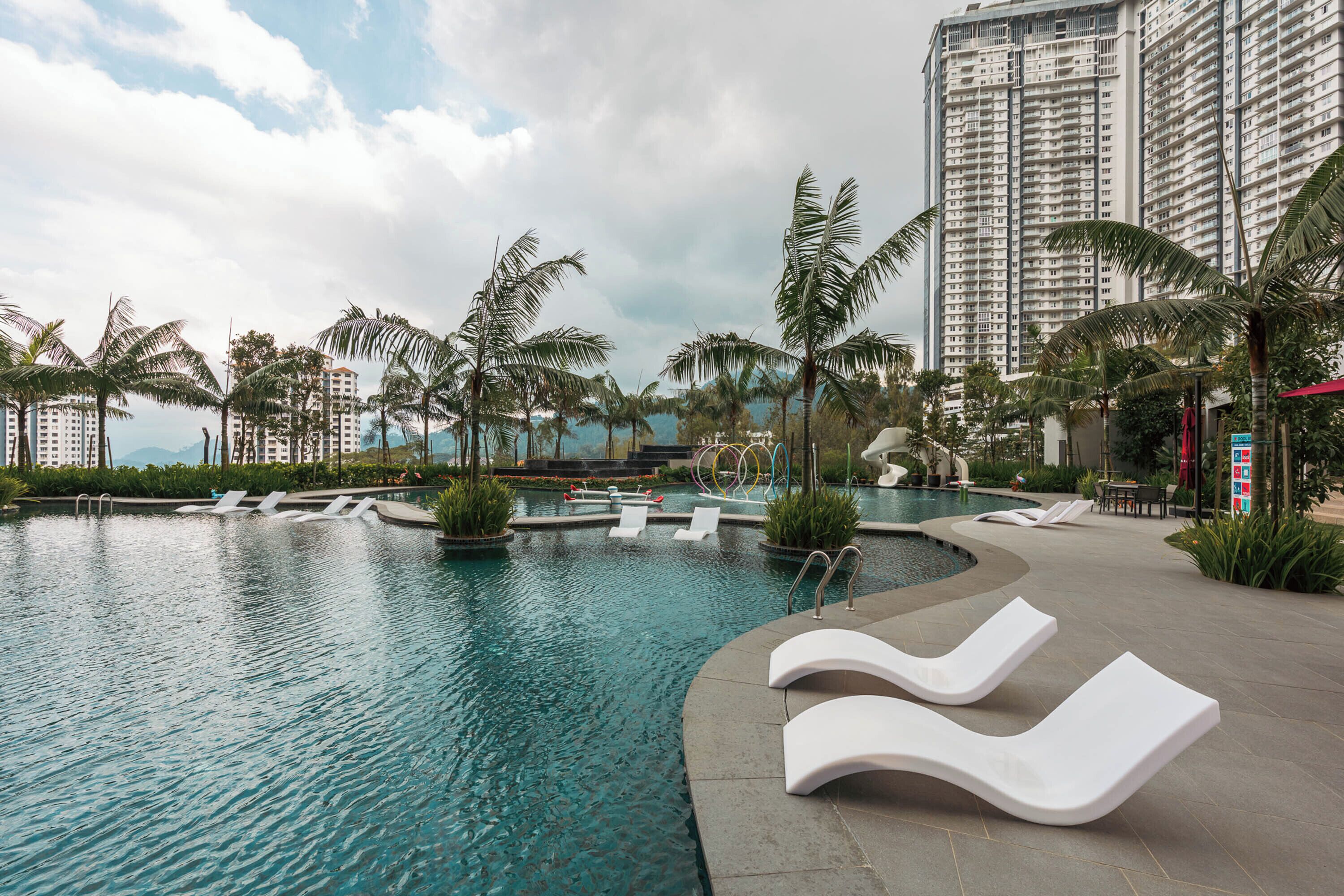 Una piscina con sillones de descanso, palmeras y edificios altos al fondo.
