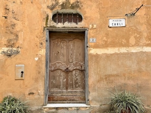 Eine verwitterte Gebäudefassade mit einer großen Holztür, einem kleinen Fenster mit Metallgitter und einem Schild mit der Aufschrift 'Piazza Carli'.