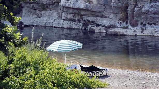 En strand med ett randigt parasoll, en stol och klippiga klippor i bakgrunden.