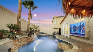 A swimming pool with a waterfall feature, surrounded by palm trees and a brick wall.