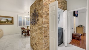 A room with a stone wall, a dining area with a table and chairs, a TV mounted on the wall, and a door leading to another room.