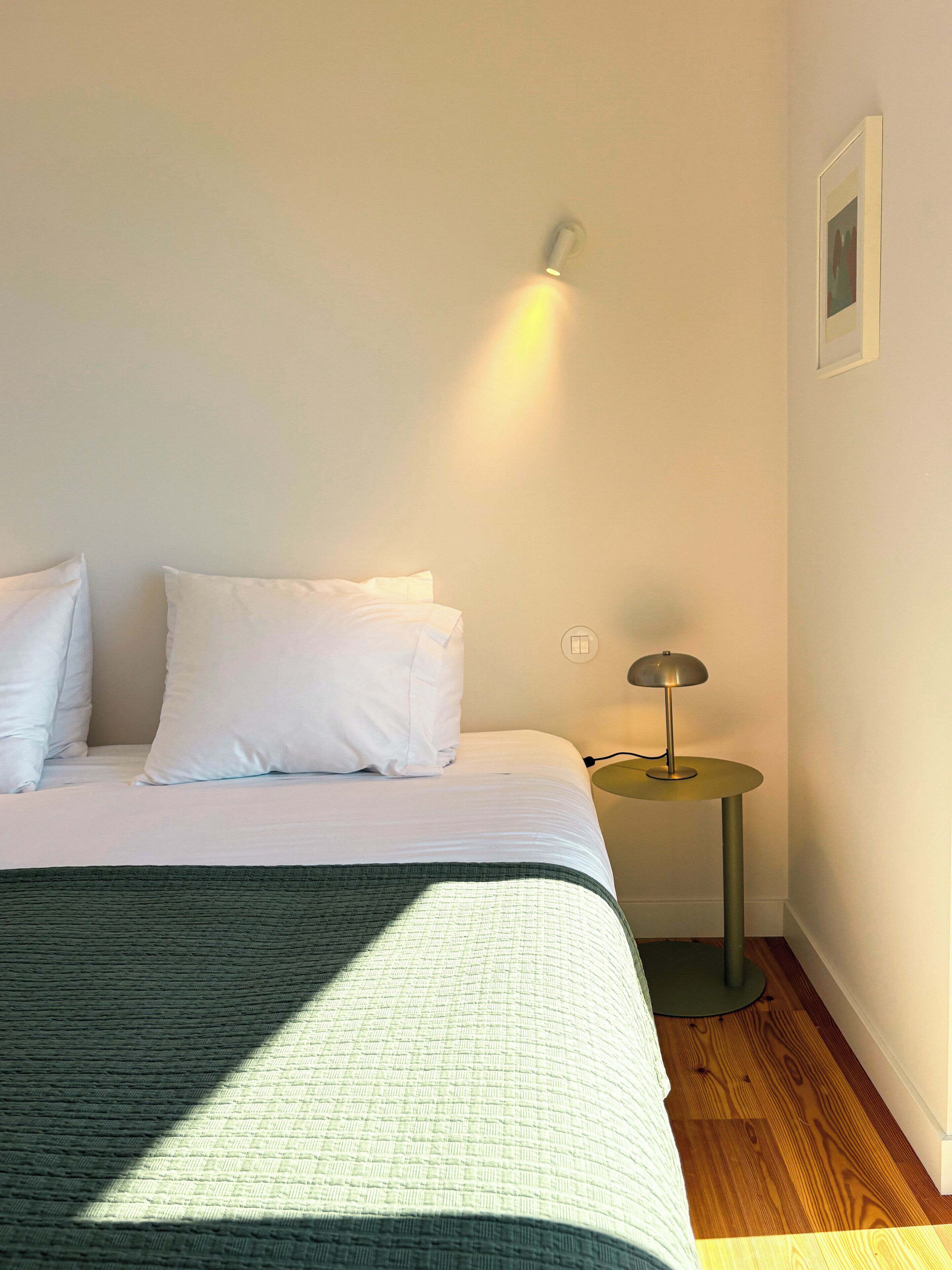 Een net opgemaakt bed met witte lakens, een klein nachtkastje met een lamp, en een lijst met een foto aan de muur.