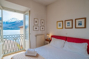 A bedroom with a bed, a nightstand, a lamp, and a view of a mountain and lake through a balcony. - Charming 170 sqm apartment, perfect for 6 people, located on the second floor of a building with an elevator (not wheelchair accessible).The property is located in the heart of Dongo (in the restricted traffic zone), a renowned town on the western shore  (Dongo)