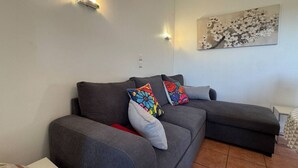 A grey sofa with colorful pillows, a wall-mounted light, and a floral painting.