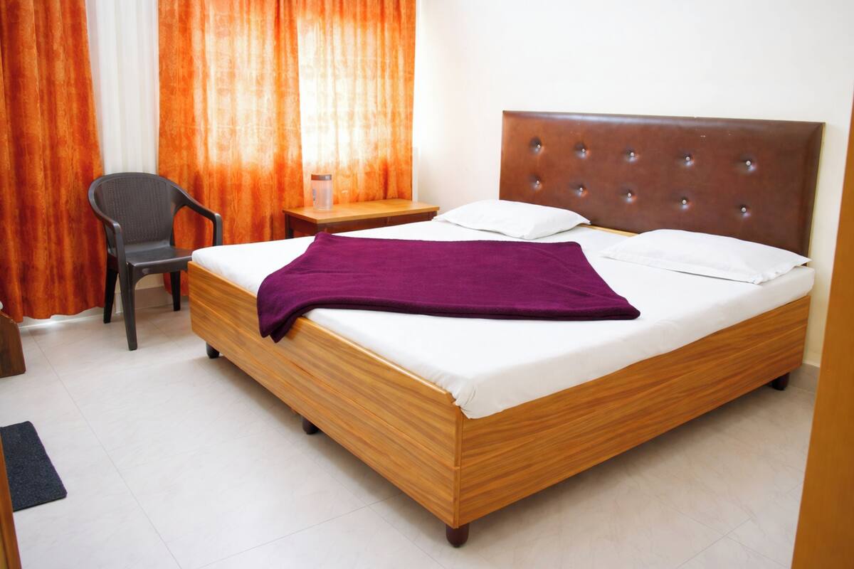 A single bed with a wooden frame and a tufted headboard, a bedside table with a lamp, a chair, and a window with orange curtains.