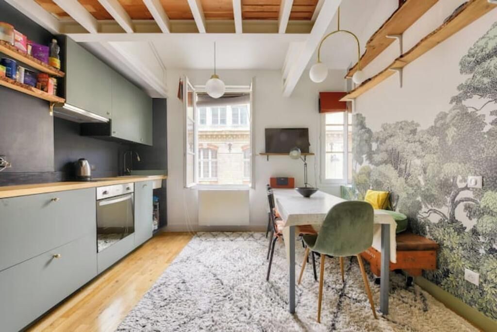Une cuisine avec des meubles de rangement en bois, une table à manger avec des chaises, un tapis et un mur orné d’une fresque représentant un arbre.