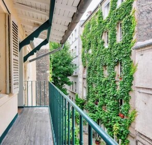 Un couloir étroit avec une rambarde métallique verte, un plancher en bois et un bâtiment recouvert de lierre.