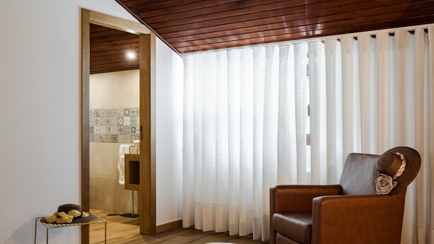 A hotel room with a bed, a brown leather chair, a small table with fruits, and a bathroom visible through a wooden door. - Holiday Home 'Jasmim' with Mountain View, Wi-Fi and Air Conditioning (Guimarães)