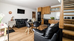 A modern living room with a black leather sofa, dining table, and chairs, a kitchen with wooden cabinets, and a TV mounted on the wall. - Holiday Home 'Jasmim' with Mountain View, Wi-Fi and Air Conditioning (Guimarães)