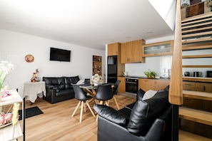 Een moderne woonkamer met een zwarte leren bank, een eettafel met stoelen, een keuken met houten kastjes en een aan de muur bevestigde televisie.