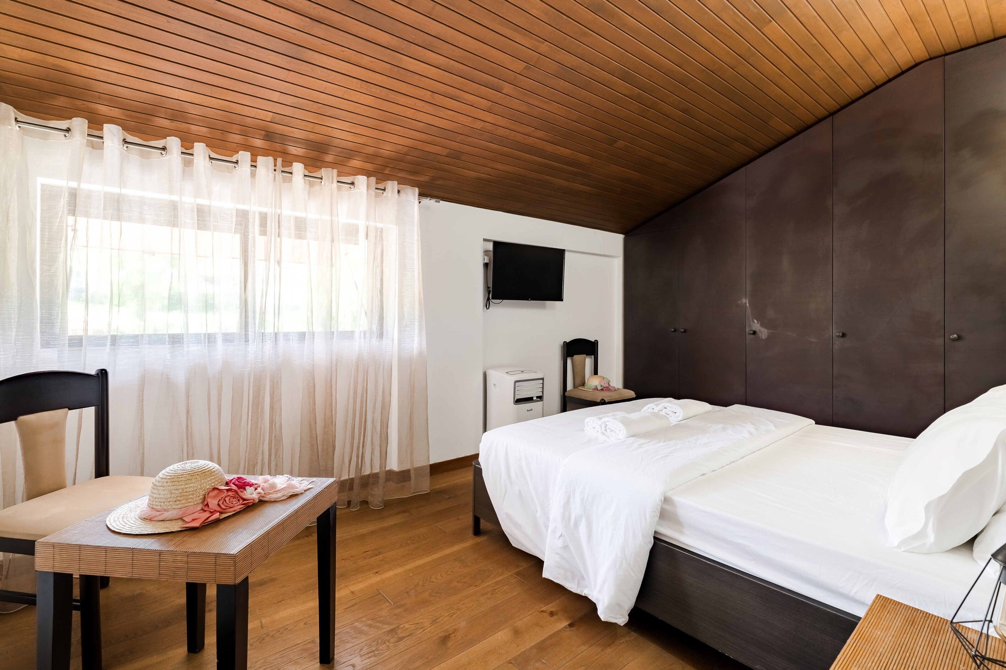 A bedroom with a wooden ceiling, a bed with white bedding, a TV on the wall, and a table with a hat and flowers.