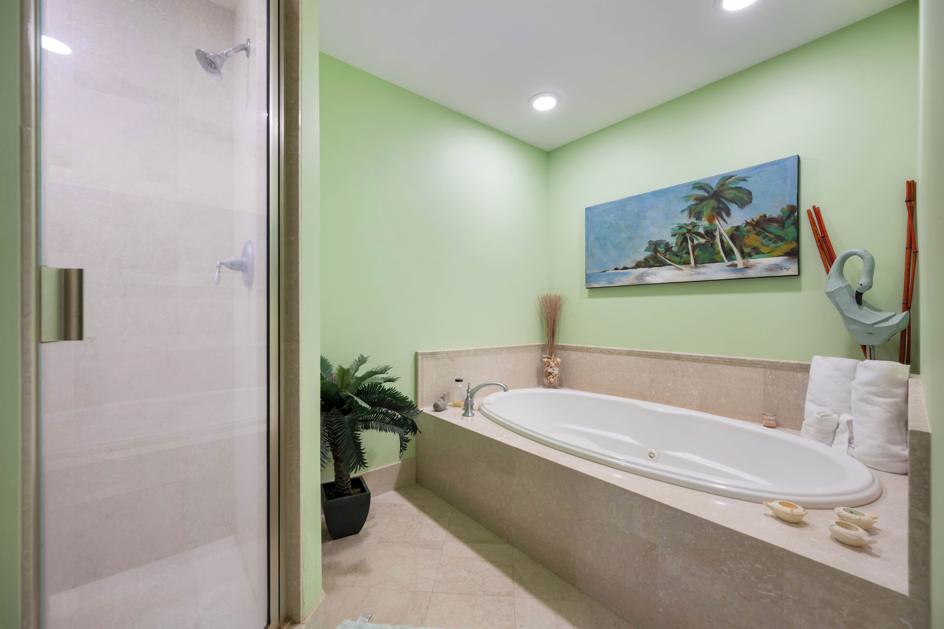A bathroom with a bathtub, a shower, a potted plant, a painting of palm trees, and a towel rack.