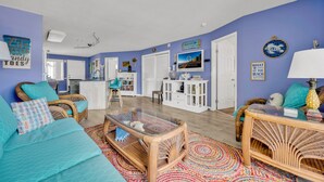 Ein Wohnzimmer mit blauer Wand, Korbmöbeln, einem Glastisch und einem farbenfrohen Teppich.