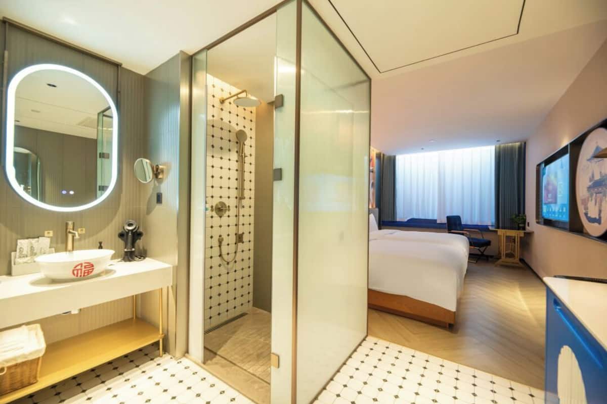 A modern hotel room with a glass-enclosed shower, a large bed, a bedside table, a TV, and a bathroom with a sink and mirror.