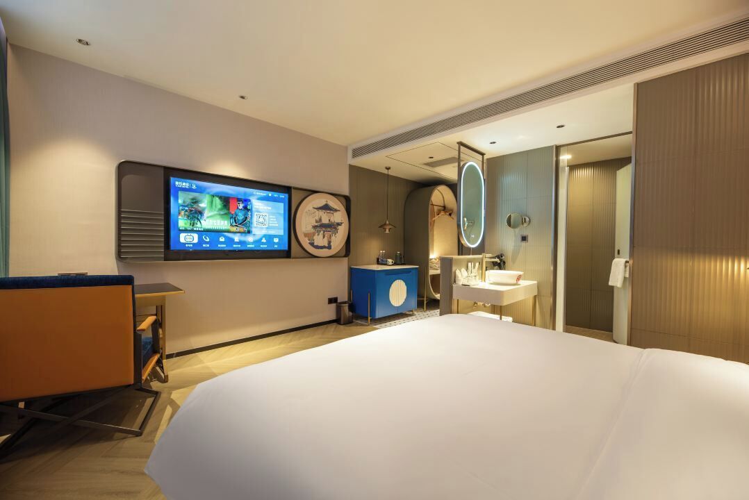 A hotel room with a bed, a TV mounted on the wall, a bathroom with a mirror, and a desk with a chair.