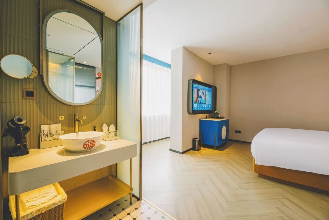 Habitación de hotel con una cama grande, un espejo redondo, un lavamanos, un televisor y un escritorio pequeño.