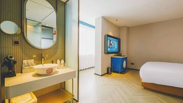 Habitación de hotel con una cama grande, un espejo redondo, un lavamanos, un televisor y un escritorio pequeño.