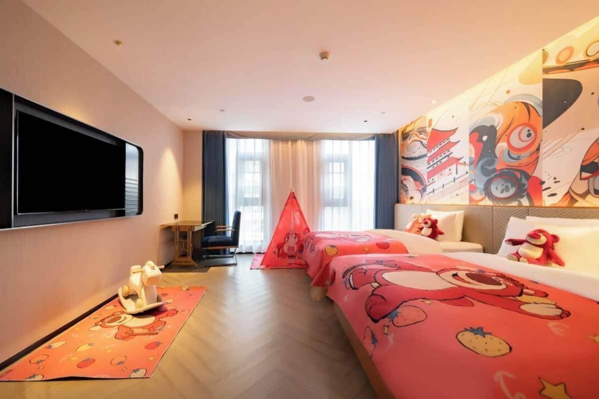 A hotel room with a large flat-screen TV, a bed with cartoon-themed bedding, a red tent-like structure, and a wall with colorful artwork.