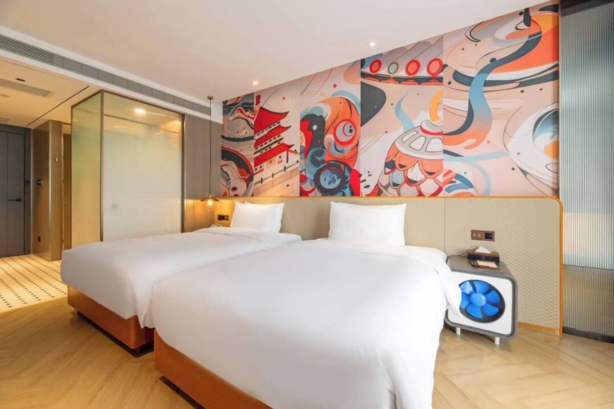 A modern hotel room with two beds, a large abstract mural, a small table with a fan, and a sliding glass door leading to a balcony.
