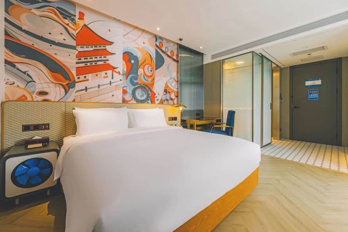 A hotel room with a large bed, a wall with a colorful mural, a small table, and a chair.