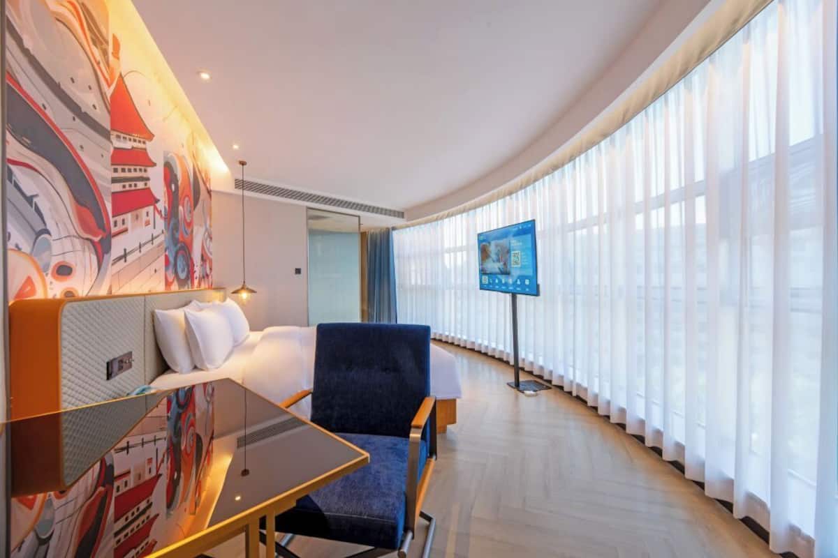 A hotel room with a bed, a desk, a chair, a TV, and large windows.