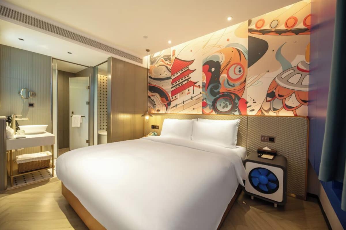 A hotel room with a large bed, a bedside table with a fan, a bathroom with a sink and mirror, and a wall with colorful artwork.