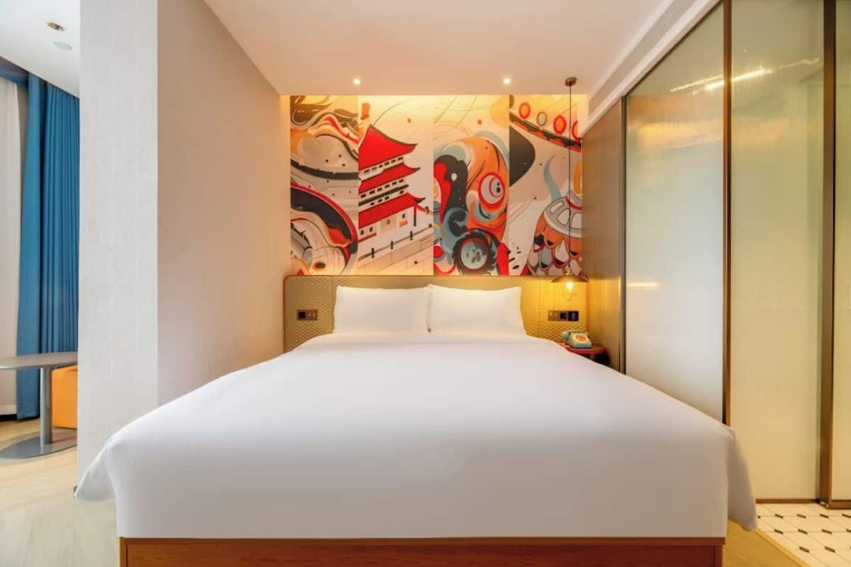 A modern hotel room with a large bed, a wall-mounted artwork, a bedside table with a lamp, and a sliding door.