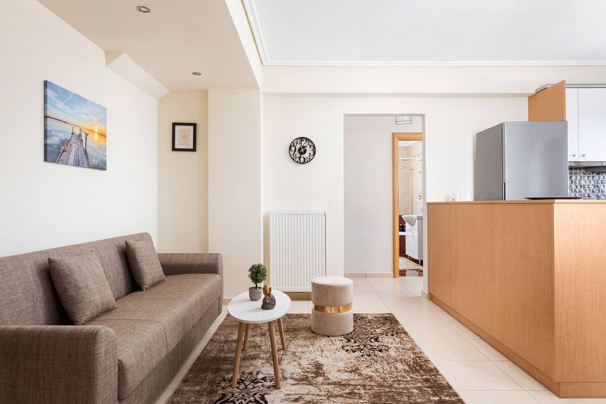 A modern living room with a sofa, a small round table, a clock, and a view into the kitchen.