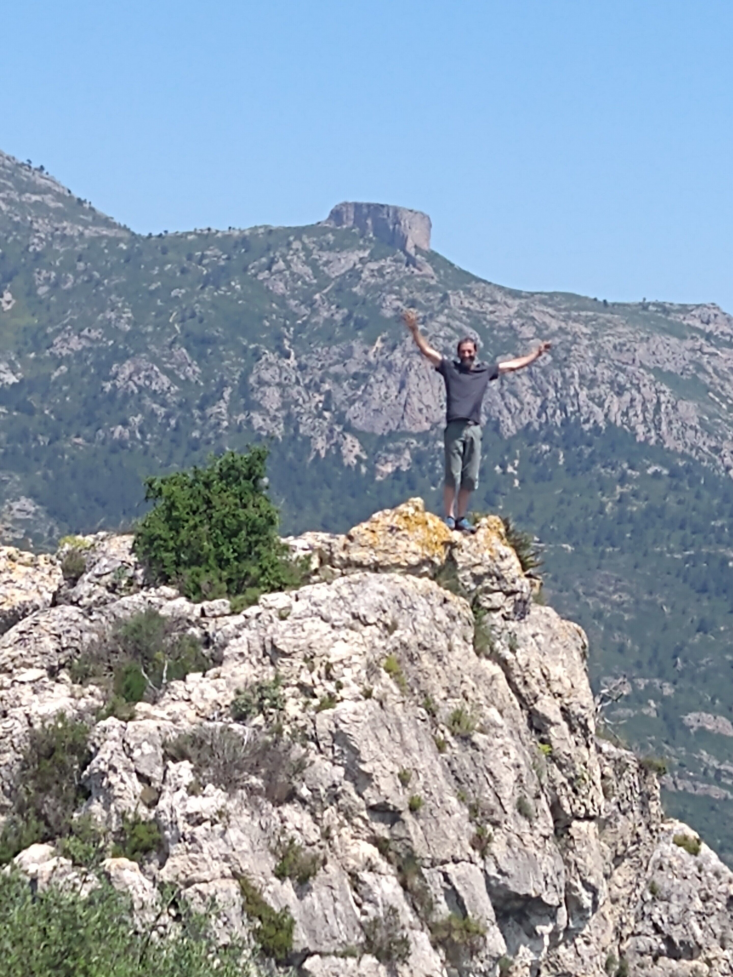 Una persona de pie sobre un afloramiento rocoso con los brazos levantados, contemplando un paisaje montañoso.