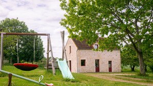 Ein rustikales Steinhaus mit roter Tür, Schaukel und Rutsche auf einer grünen Wiese mit Bäumen.