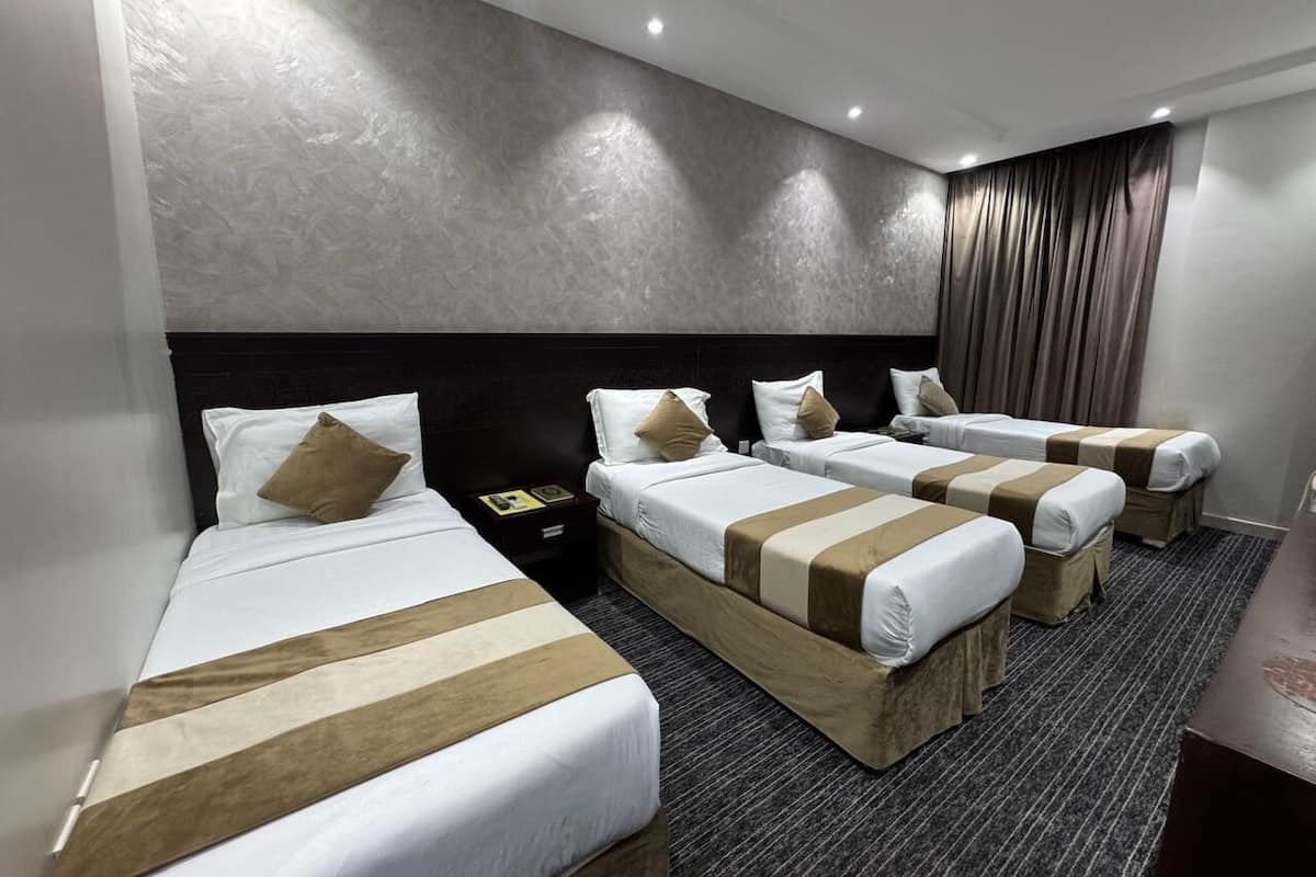 Habitación de hotel con cuatro camas, cabecera oscura, suelo alfombrado y una mesita de noche pequeña con lámpara.