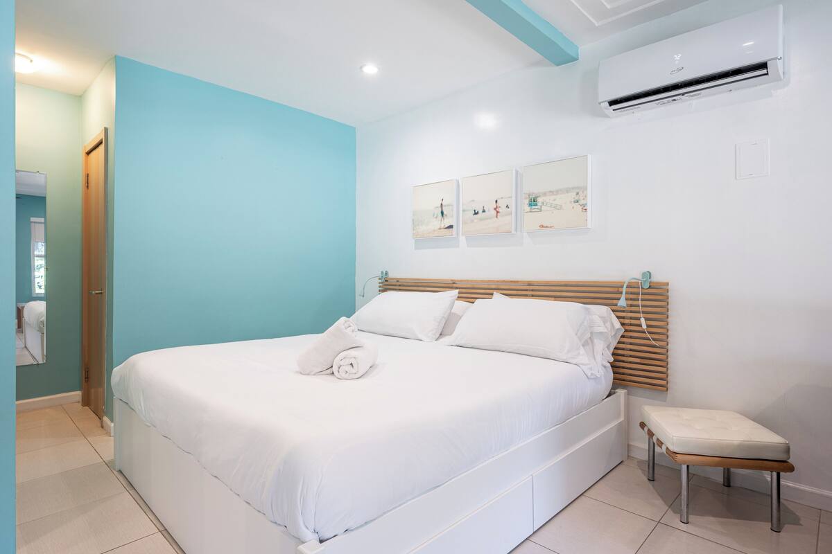 Una cama bien hecha con sábanas blancas y una toalla enrollada, un banco de madera a un lado y un aire acondicionado de pared.
