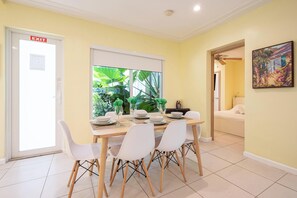 Uma sala de jantar com uma mesa posta para quatro pessoas, cadeiras brancas, uma janela com vista para o verde, uma porta de saída e um quadro na parede.
