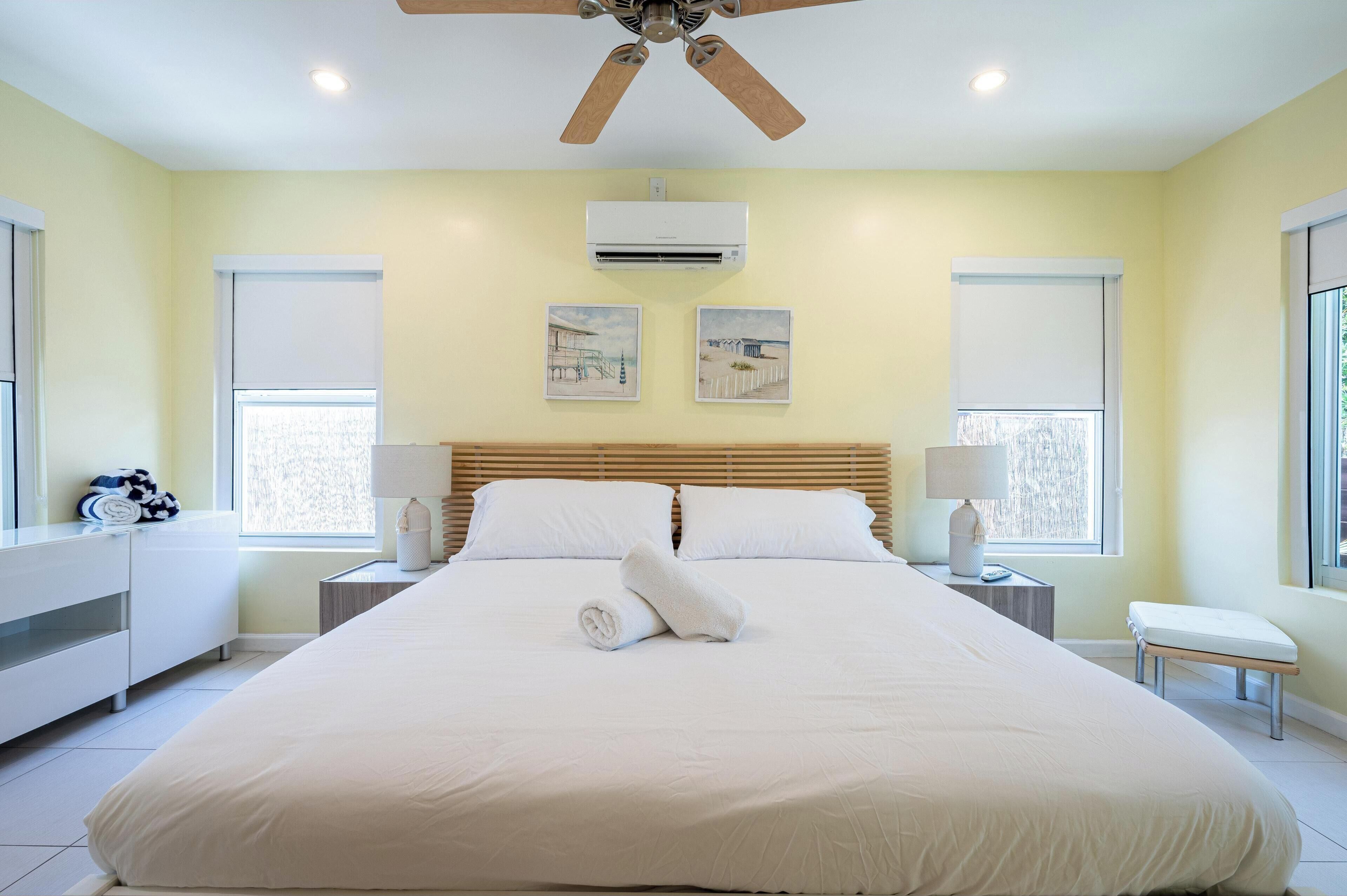 A hotel room with a bed, ceiling fan, air conditioning unit, two wall-mounted lamps, and a window with blinds.
