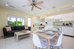 Sala de estar moderna com sofá, mesa de centro, mesa de jantar e cadeiras brancas. Há um ventilador de teto, uma cozinha com micro-ondas e uma janela com vista para o verde.