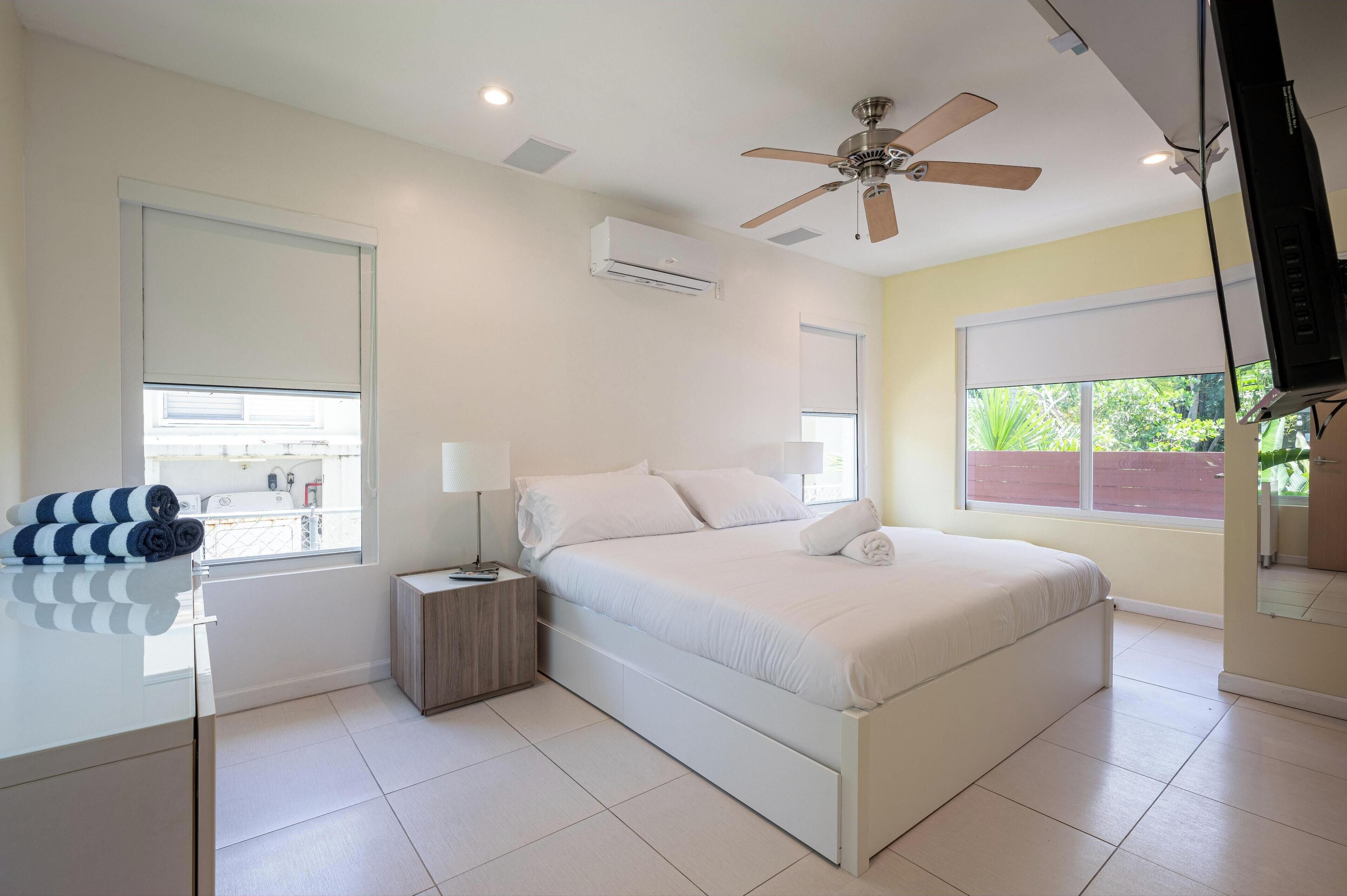 Un dormitorio con cama, mesita de noche, ventilador de techo, aire acondicionado y ventana con vistas.