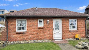 Ein Ziegelhaus mit weißer Tür, zwei Fenstern, einer kleinen Veranda und einem Kiesweg.