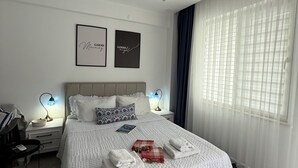Une chambre à coucher bien aménagée, avec un grand lit, des tables de chevet, une petite table d’appoint, une lampe fixée au mur et une fenêtre ornée de rideaux légers.