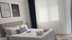 Une chambre à coucher moderne avec un lit, des tables de chevet, une petite table, une fenêtre avec des rideaux légers et deux citations encadrées accrochées au mur.