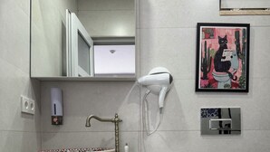 Une salle de bain avec un lavabo blanc, un grand miroir, un sèche-cheveux, des toilettes et un tableau encadré au mur.