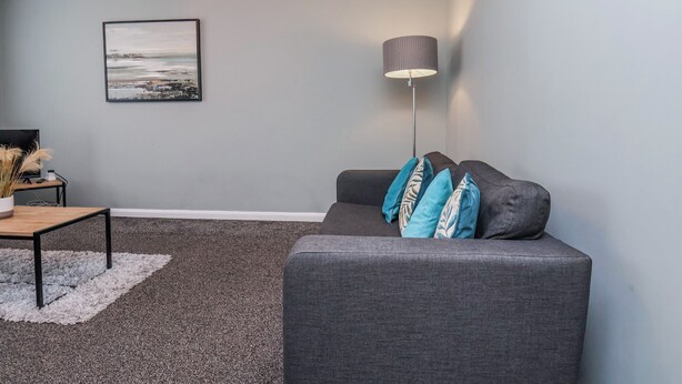 Een woonkamer met een grijze bank, een vloerlamp, een lijst met een foto aan de muur, en een klein tafeltje met een vaas.