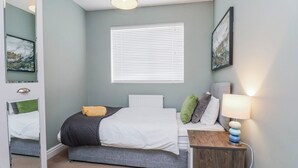 Een slaapkamer met een bed, nachtkastje, lamp, raam en kunstwerk.