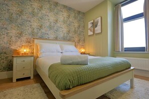A bedroom with a bed, bedside lamps, a window with curtains, and a floral wallpaper. - The Tall Trees Hotel (Amlwch)