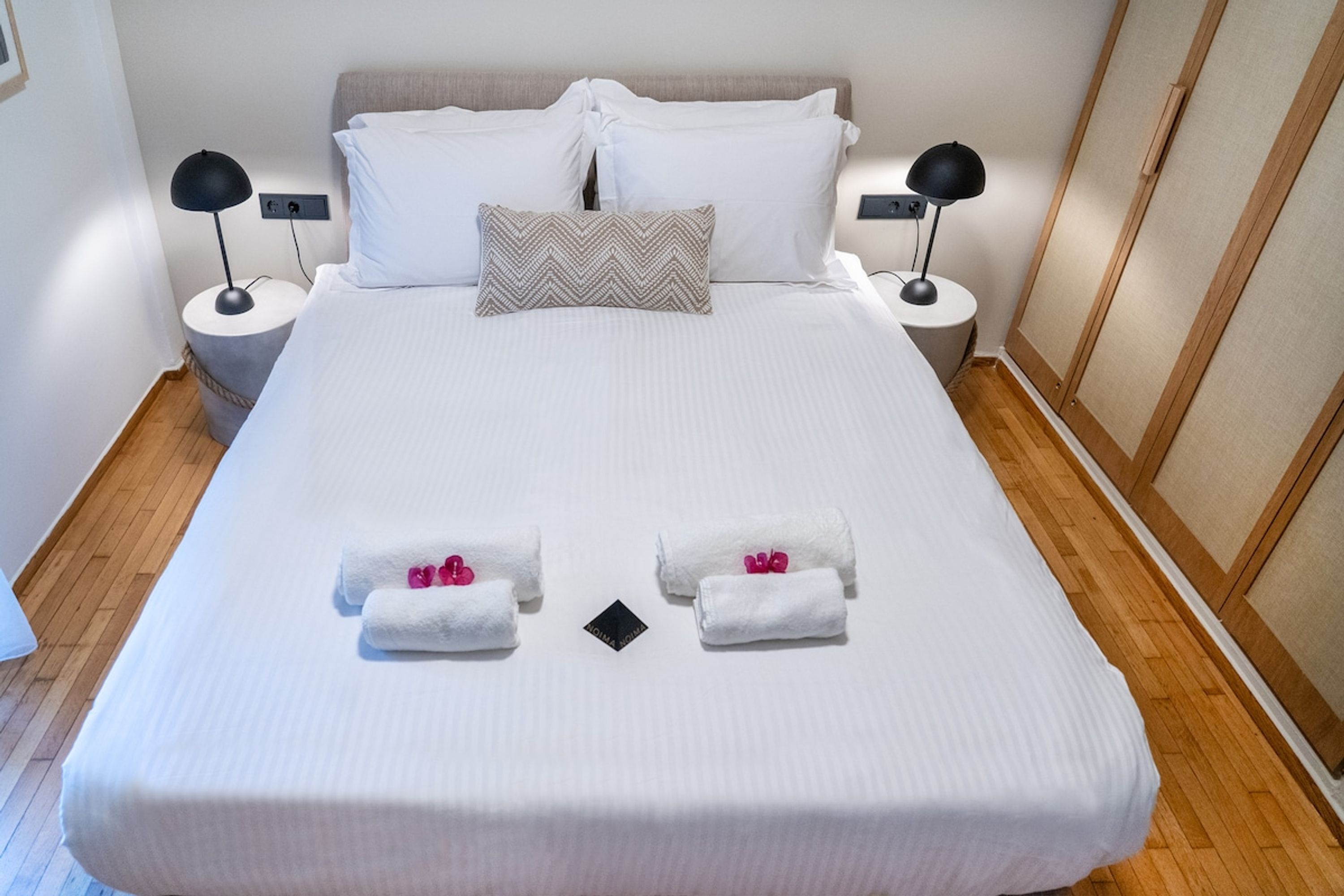 A neatly made bed with white linens, two pillows, two bedside lamps, and two towels with pink flowers.