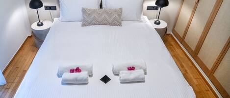 A neatly made bed with white linens, two pillows, two bedside lamps, and two towels with pink flowers.