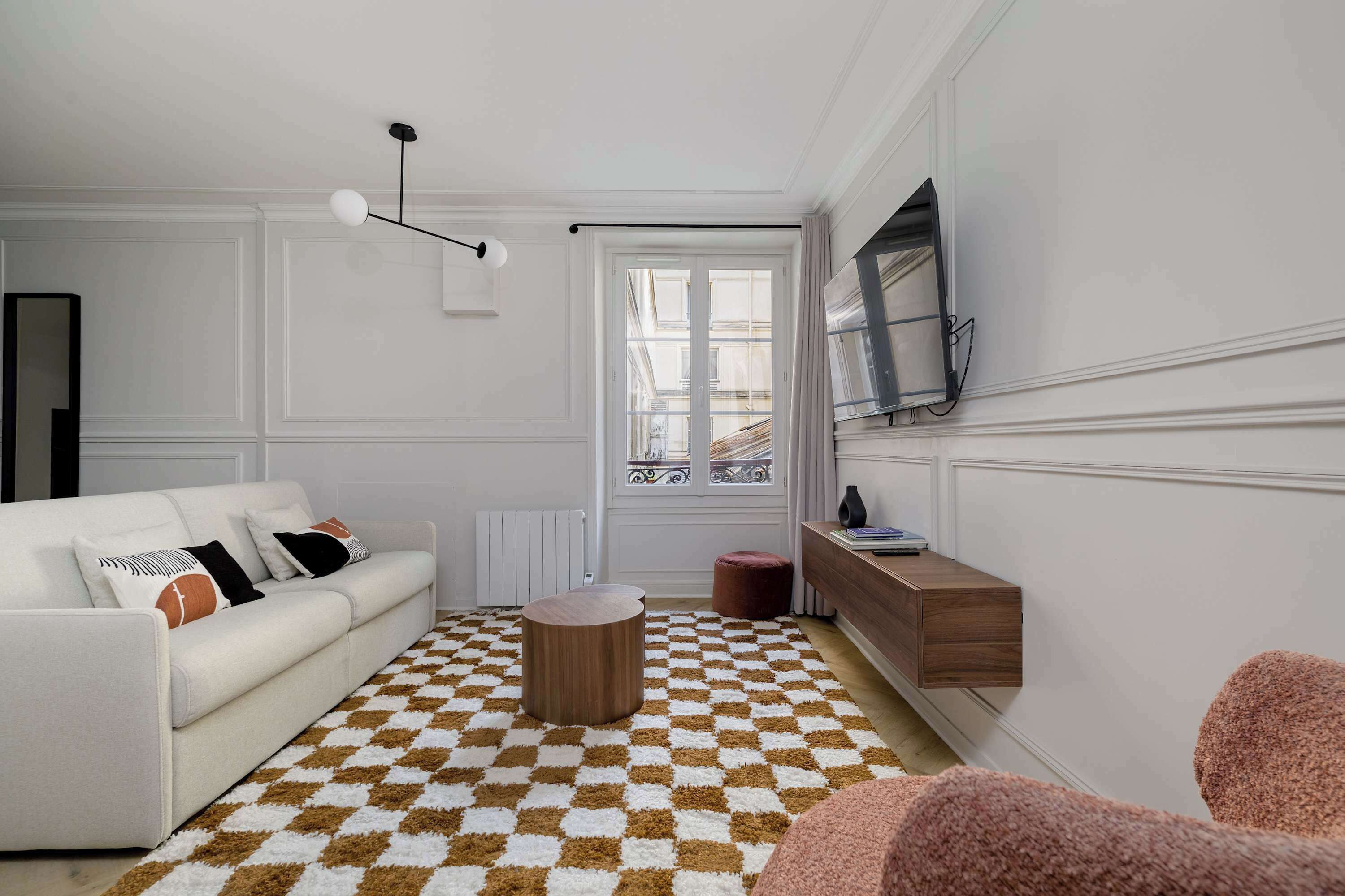 A living room with a white sofa, a round wooden coffee table, a checkered rug, a television mounted on the wall, and a window with a view of buildings.