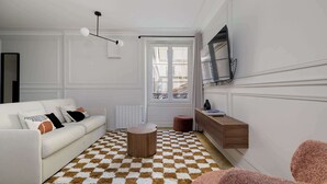 Sala de estar com sofá branco, mesa de centro redonda de madeira, tapete xadrez, televisão fixada na parede e janela com vista para prédios.