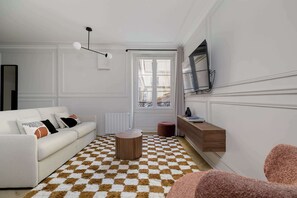 Ein Wohnzimmer mit einem weißen Sofa, einem runden Holztisch, einem karierten Teppich, einem an der Wand montierten Fernseher und einem Fenster mit Blick auf Gebäude.