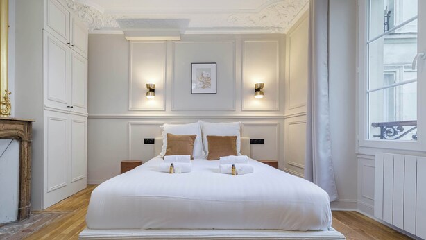 Uma cama grande com roupa de cama branca, duas mesas de cabeceira, arandelas na parede e uma obra de arte emoldurada sobre a cama.