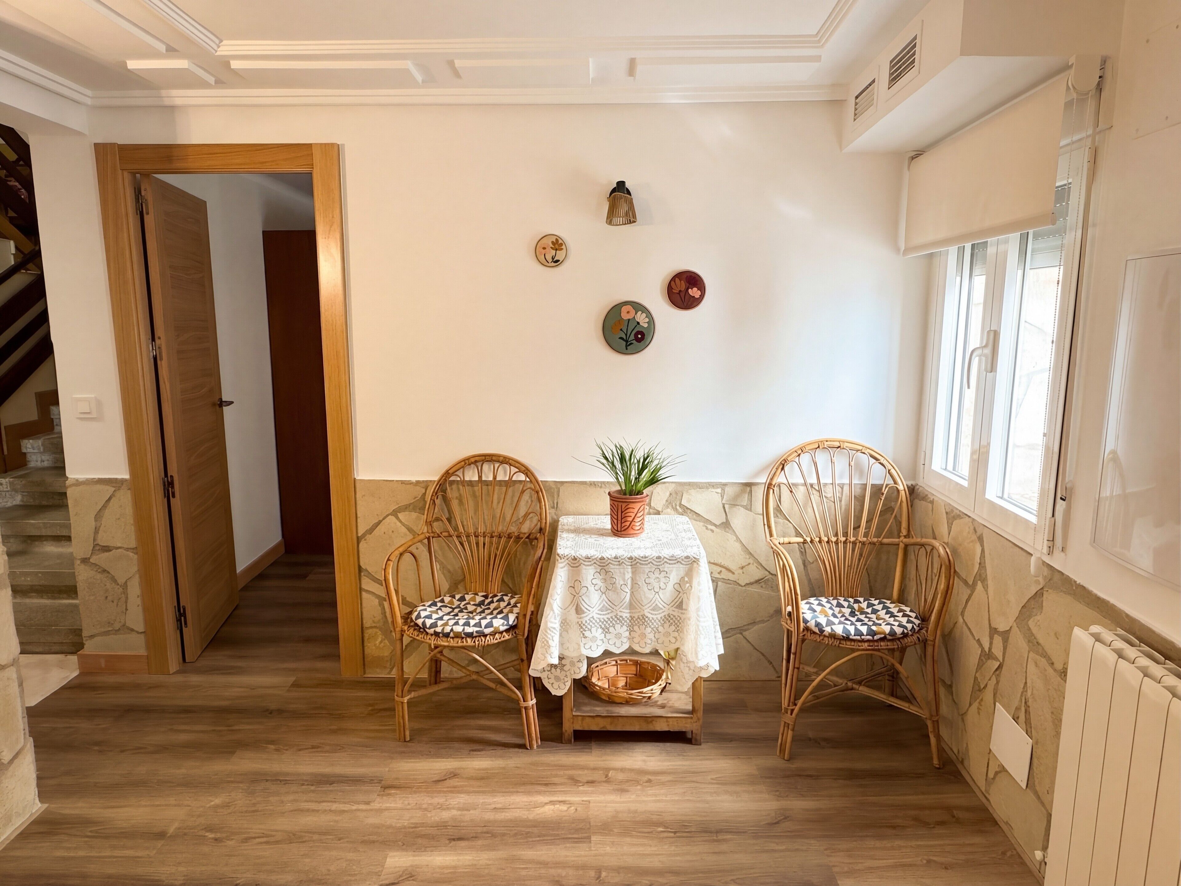 Una habitación con piso de madera, dos sillas de mimbre, una mesa con mantel de encaje, una planta en maceta y una pared de piedra.